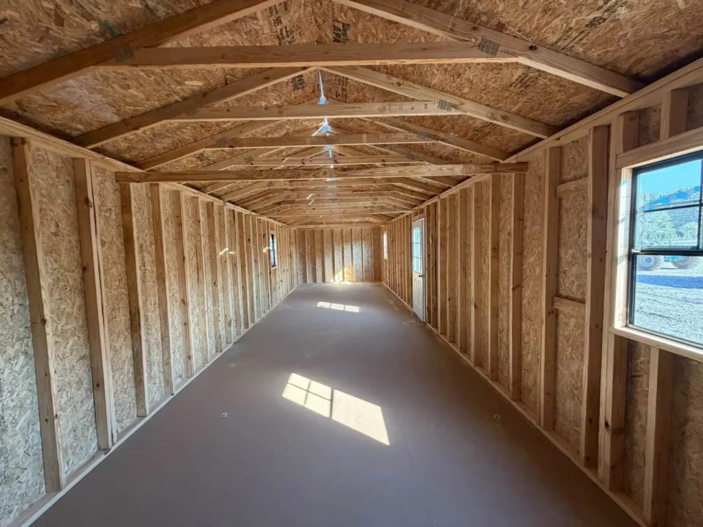 Inside an unfinished 12x40 portable building: exposed wood framing and roof trusses, windows on both sides, and a 9-lite door at the far end