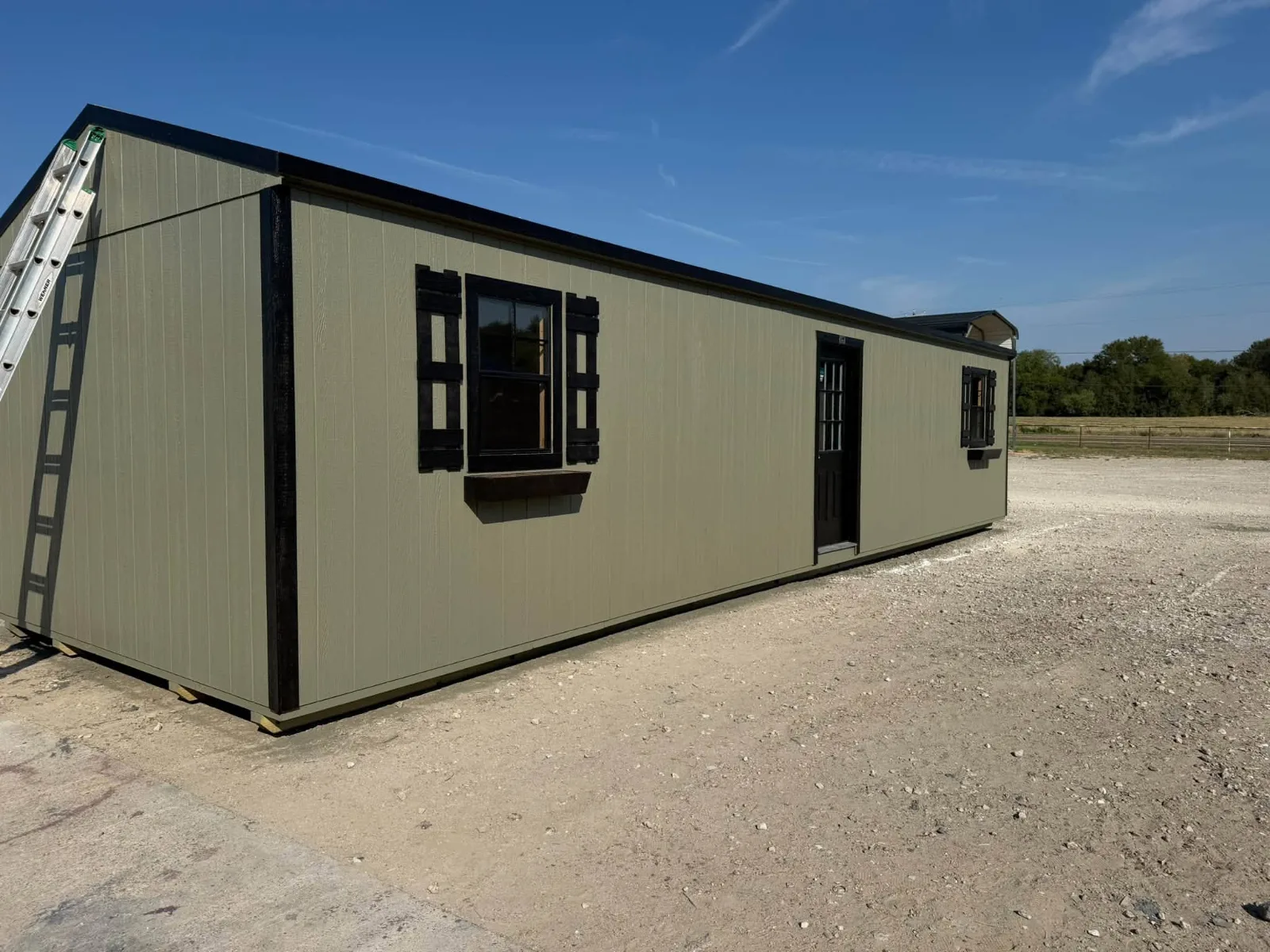 12x40 portable building with sage green siding, black trim, a black window with shutters and a flower box, and a 9-lite door, at the Corsicana yard
