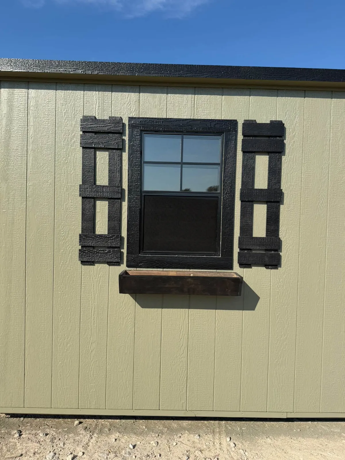 Close-up of a black-framed window with black shutters and a wooden flower box on sage green portable building siding