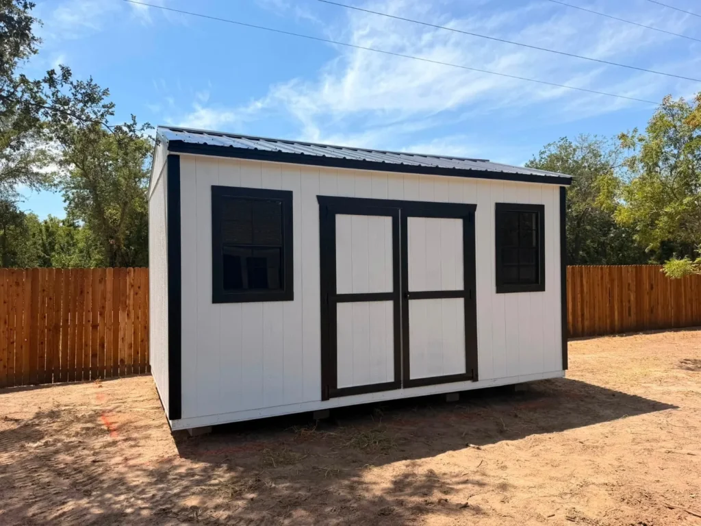 White utility shed with black trim, black-framed windows, and black and white double doors, set beside a wood privacy fence near Denton